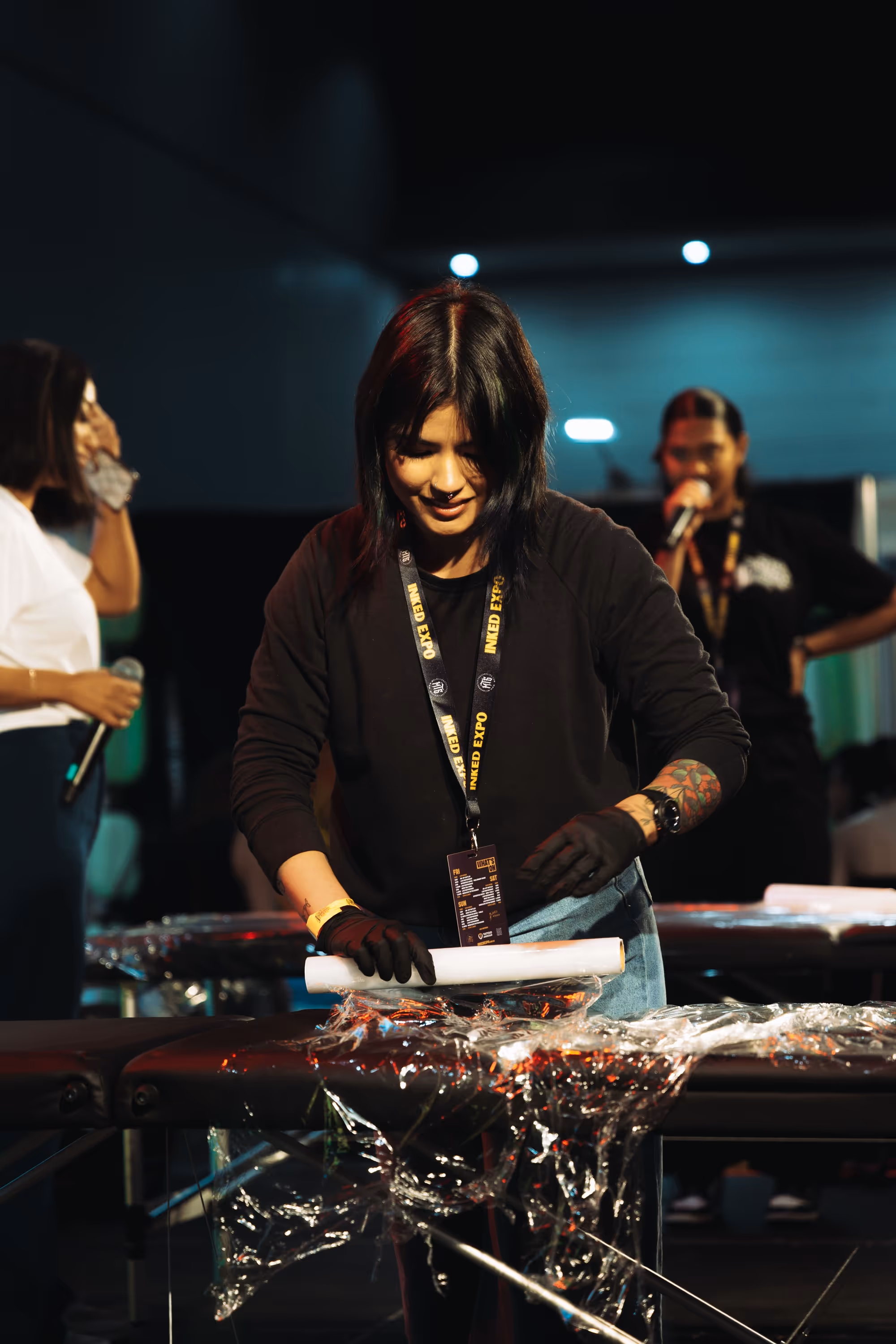Woman wearing black gloves preparing a tattoo station covered in plastic wrap.