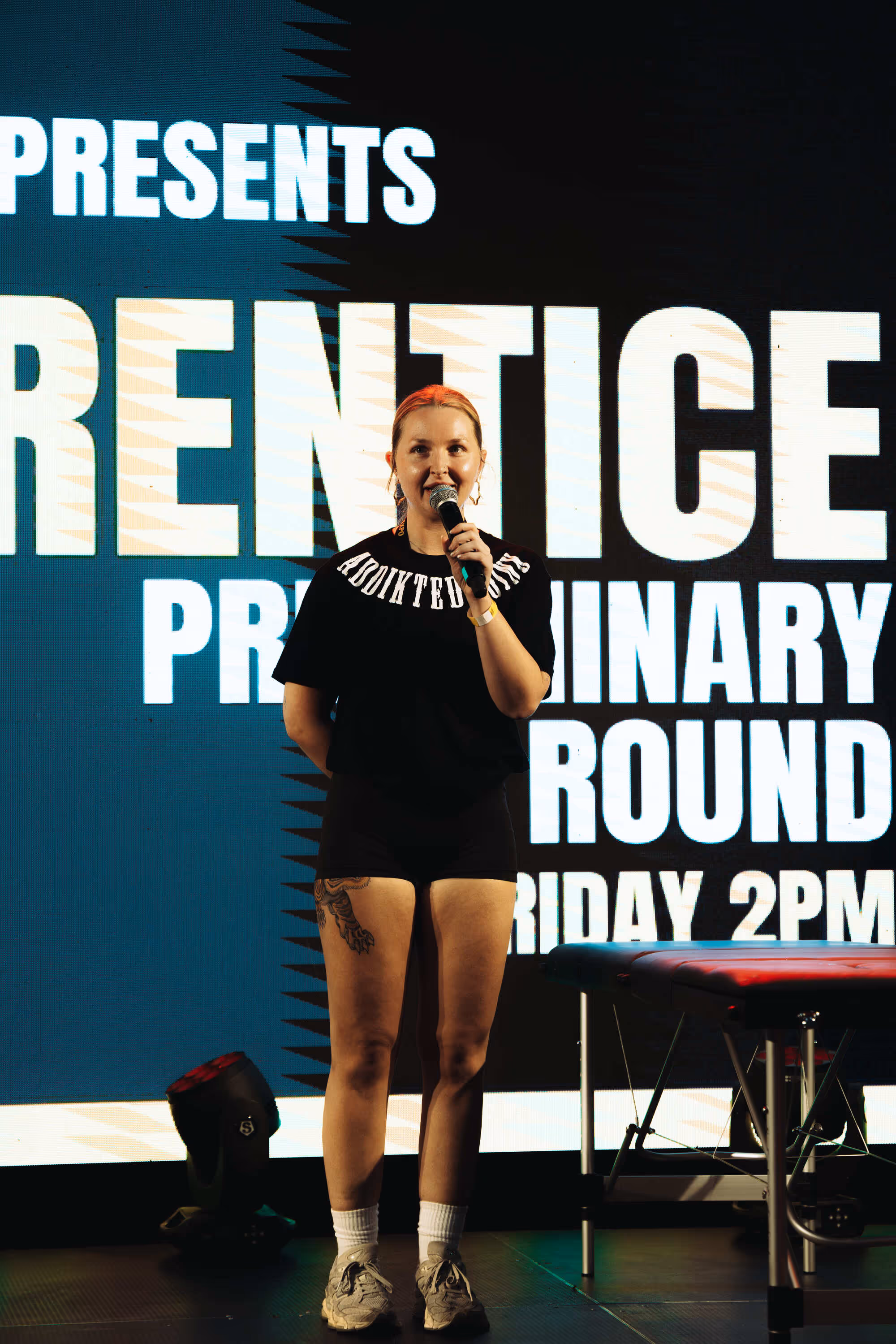 Woman with a microphone standing on stage in front of a large screen with event text.