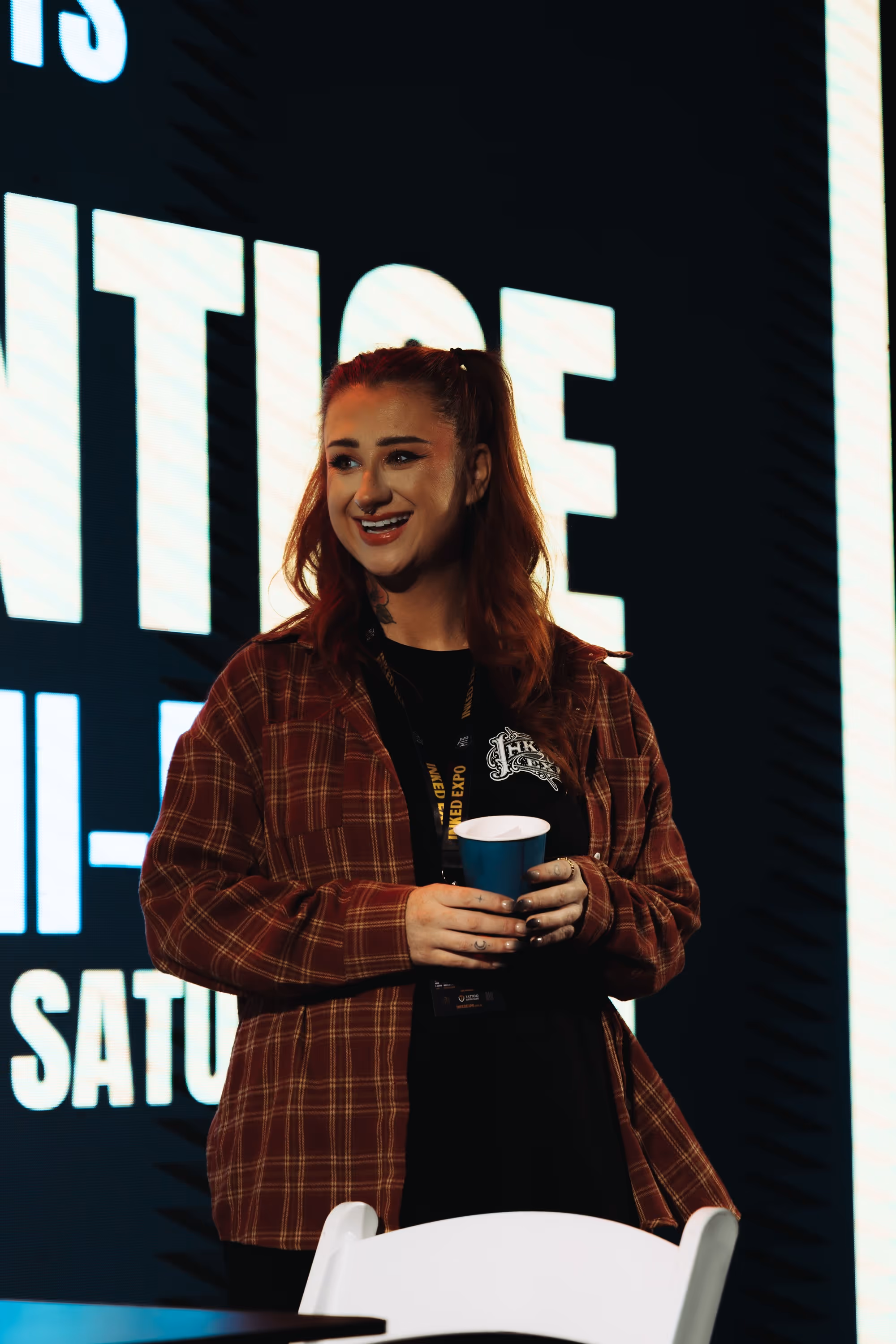 Smiling woman with red hair in a red plaid shirt holding a blue cup and standing in front of a large illuminated sign.