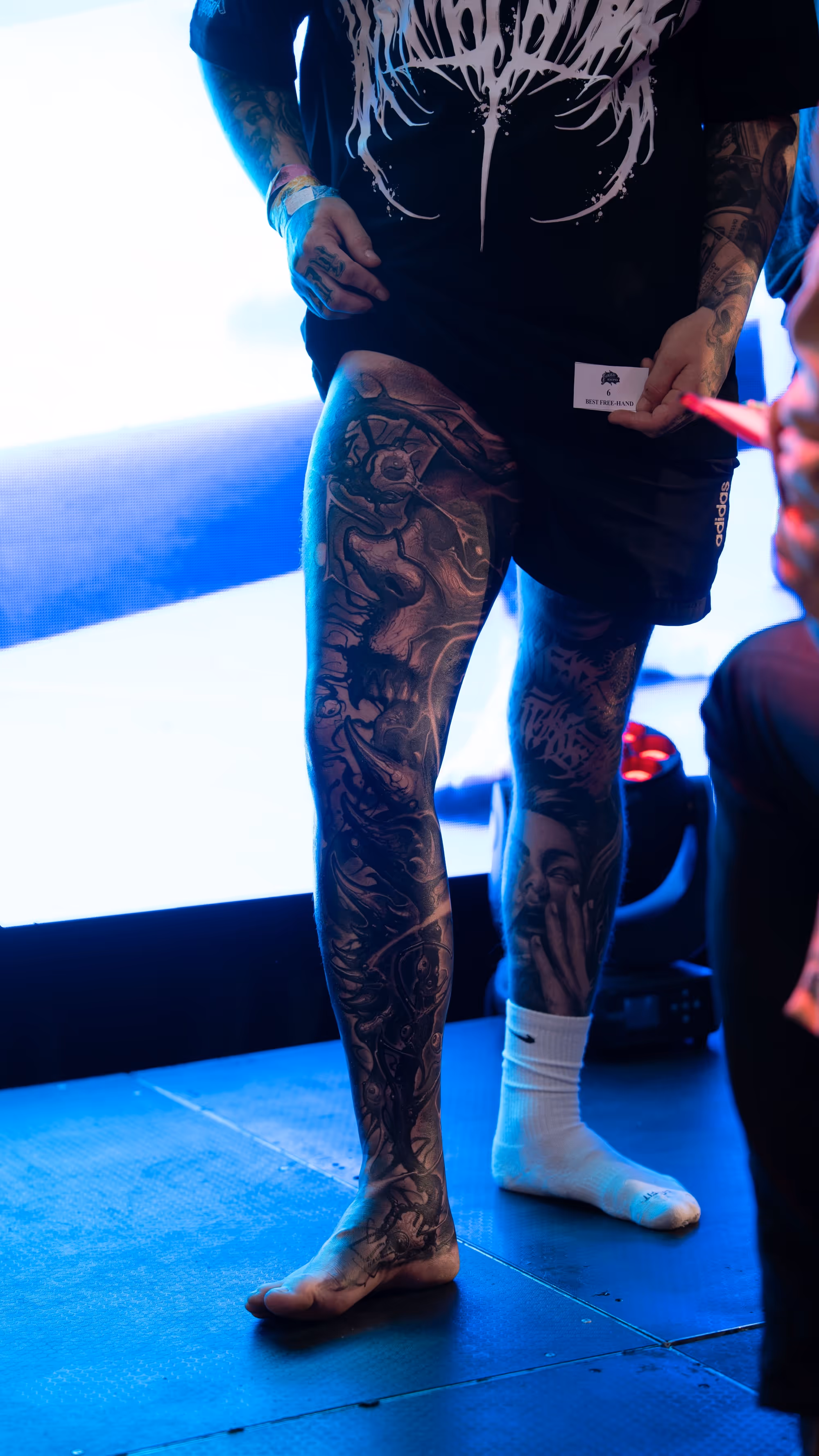 Person with detailed black and grey leg tattoos, wearing a black shirt, black shorts, one white sock, and holding a card that reads 'Best Free-Hand'.