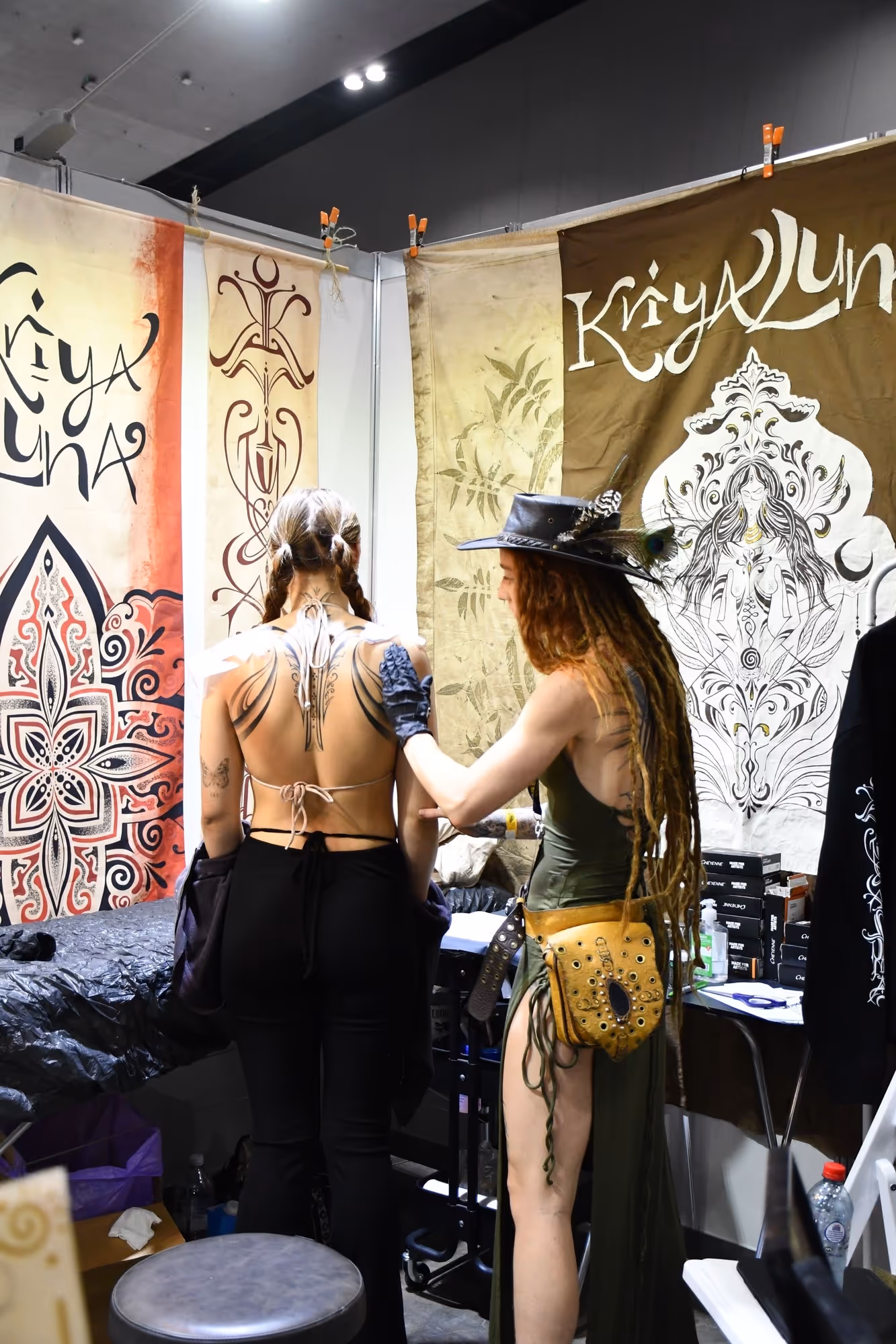 A woman with long dreadlocks wearing a hat and green dress applies a tattoo on the upper back of another woman with braided hair.