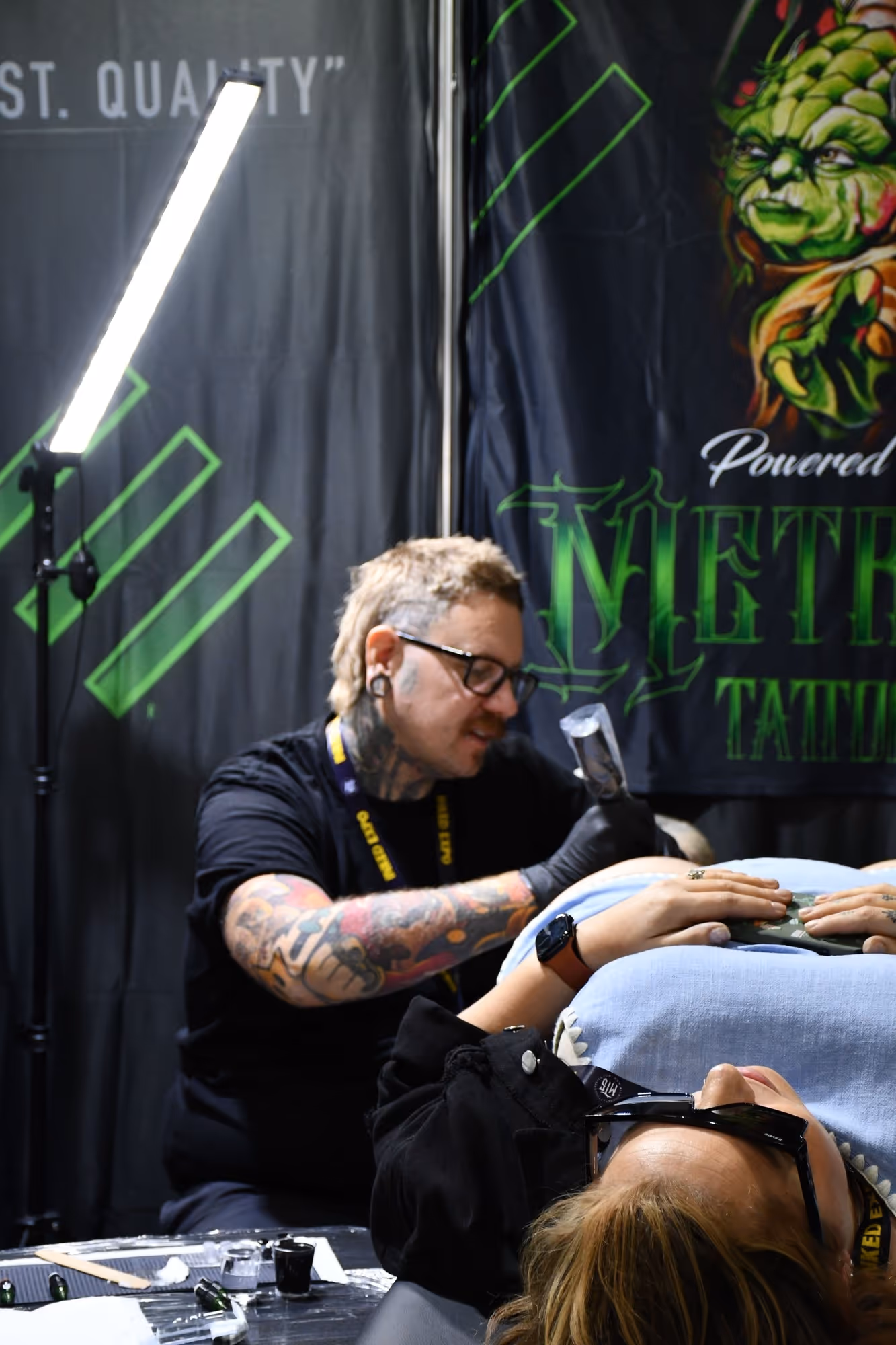Tattoo artist with glasses and colorful sleeve tattoos working on a client lying down, with a bright light and Metro Tattoo banner in the background.