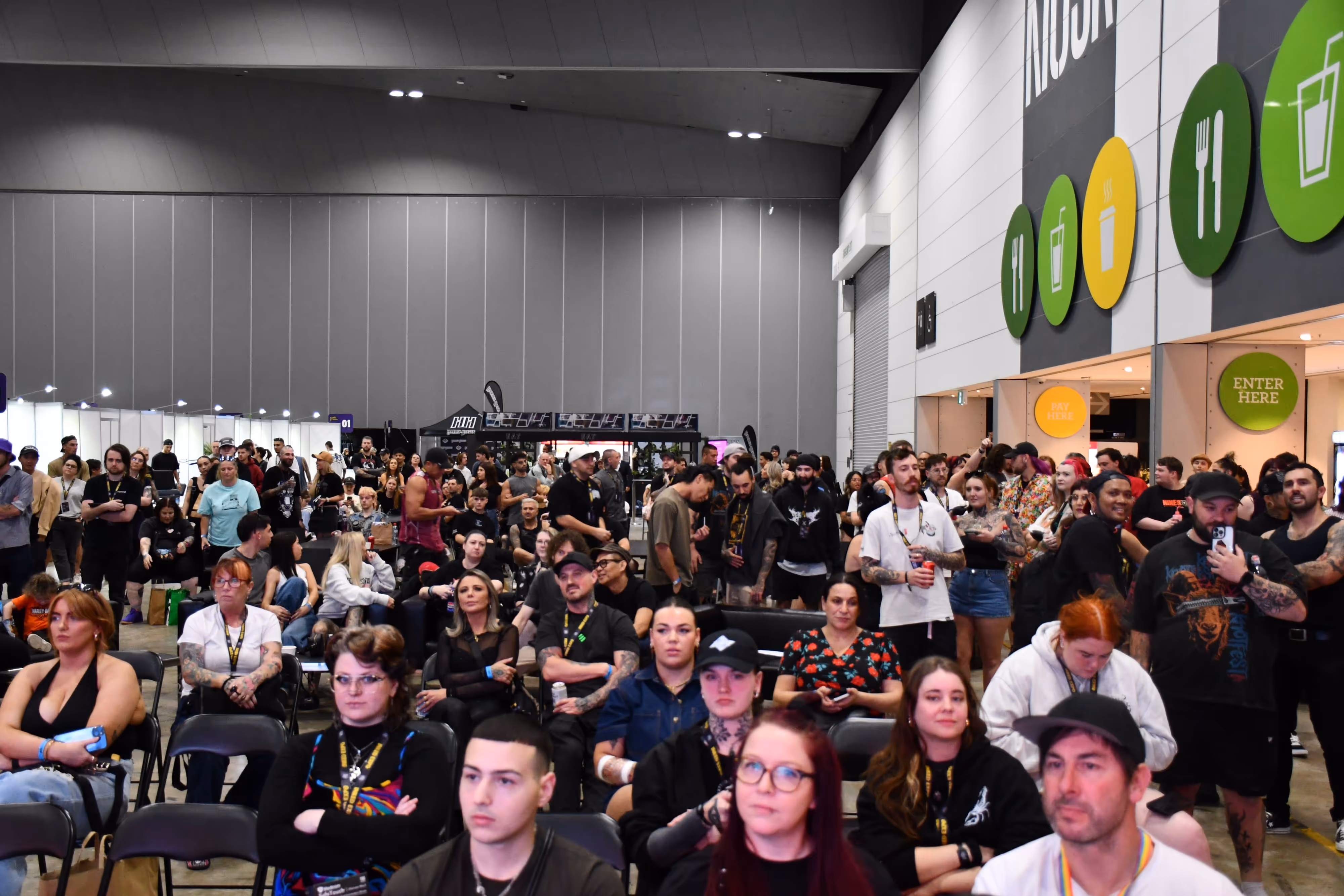 Crowd of diverse people seated and standing indoors at a convention or event with food signage visible on the wall.