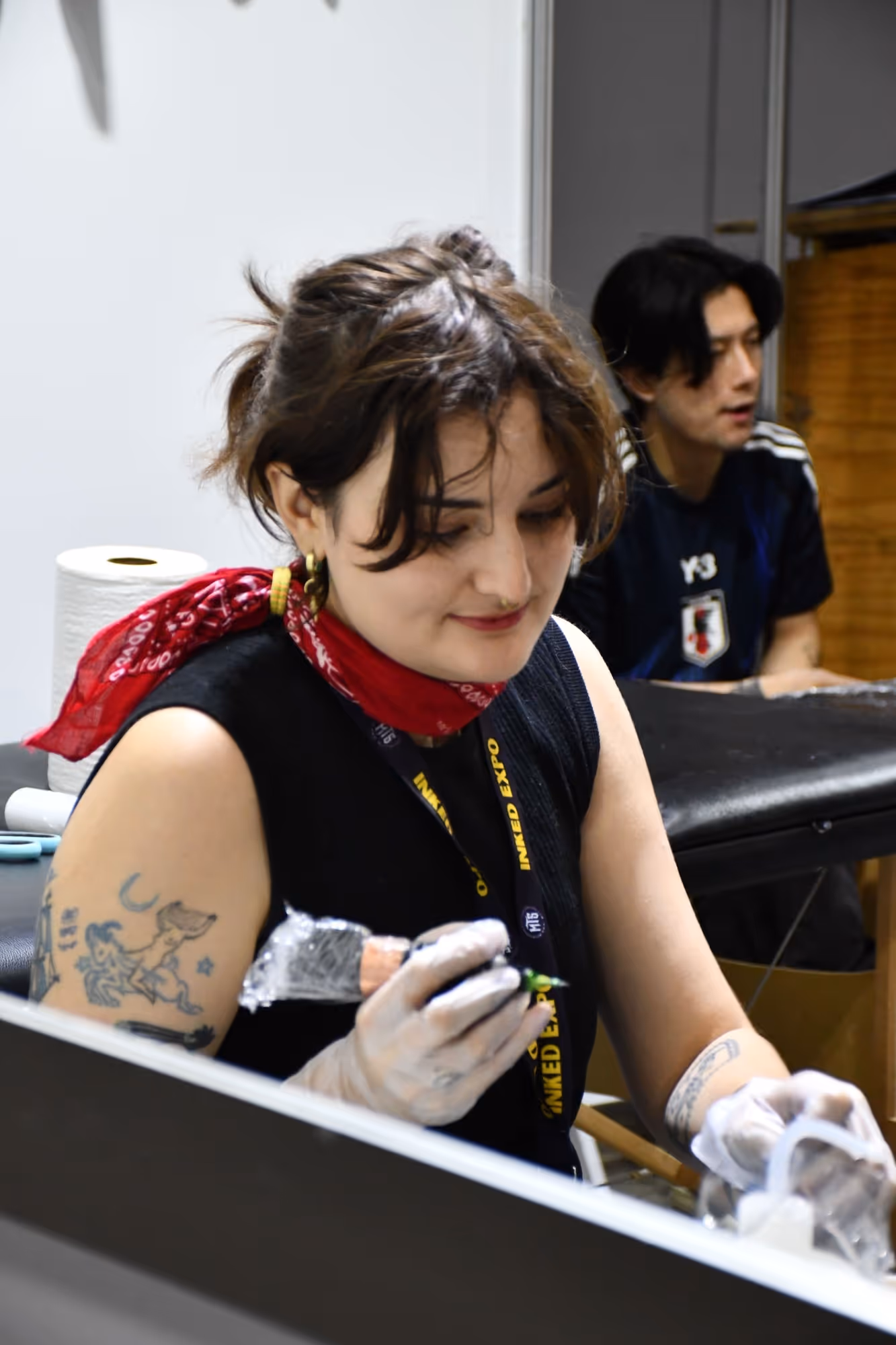 Tattoo artist wearing gloves and a red bandana around her neck, focusing on her work inside a studio.