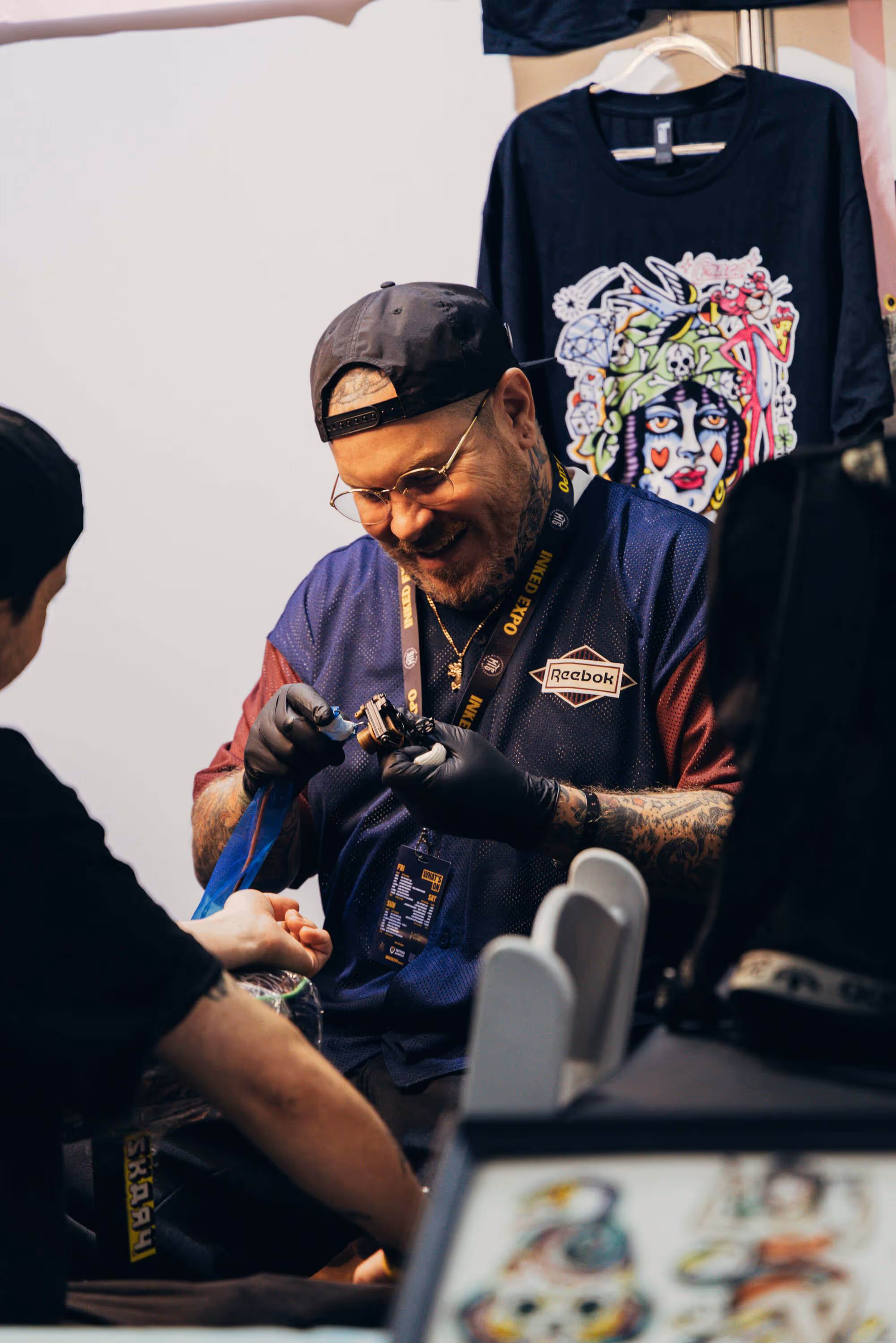 Tattoo artist wearing a cap and glasses, smiling while tattooing a client's arm at a convention booth.