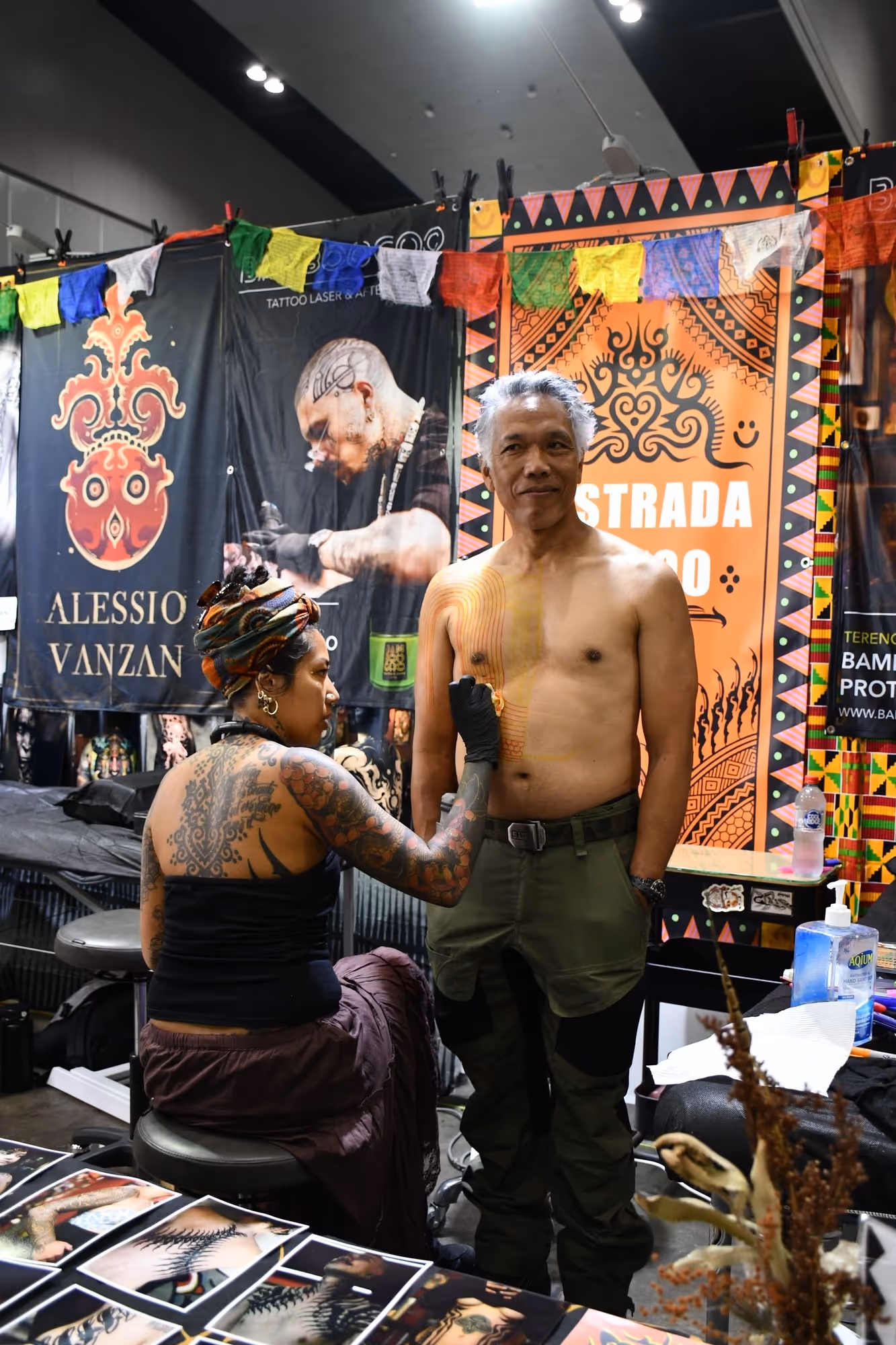 Tattoo artist with sleeve tattoos working on chest tattoo of a smiling shirtless man in a tattoo convention.