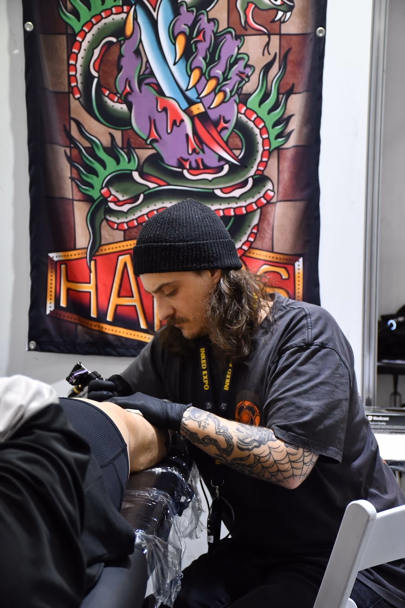 Tattoo artist with a beanie and gloves tattooing a person's arm in a studio with a colorful snake and dagger banner in the background.