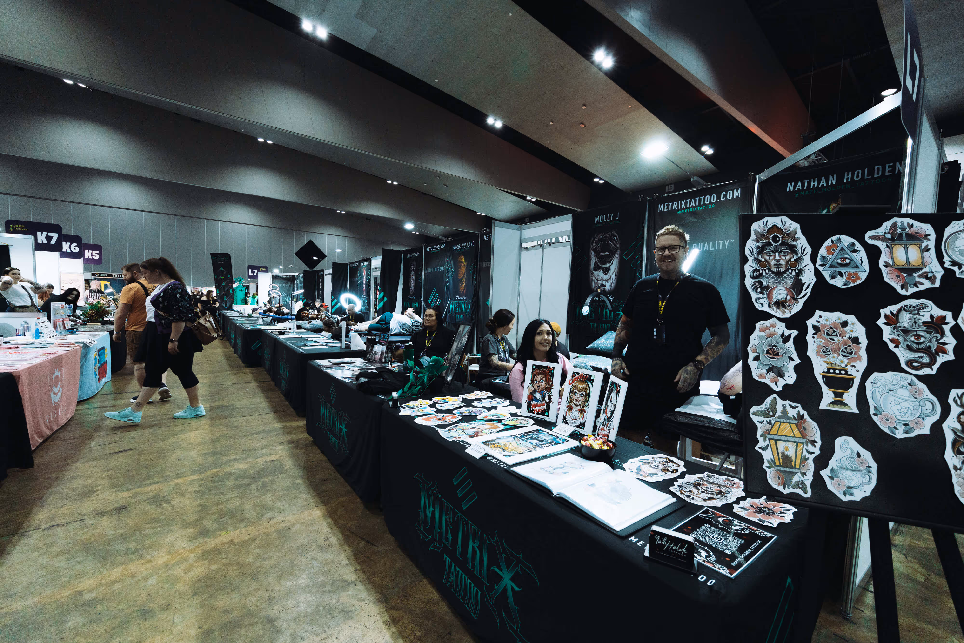 Tattoo convention booth with artists showcasing tattoo designs and artwork on tables and boards.