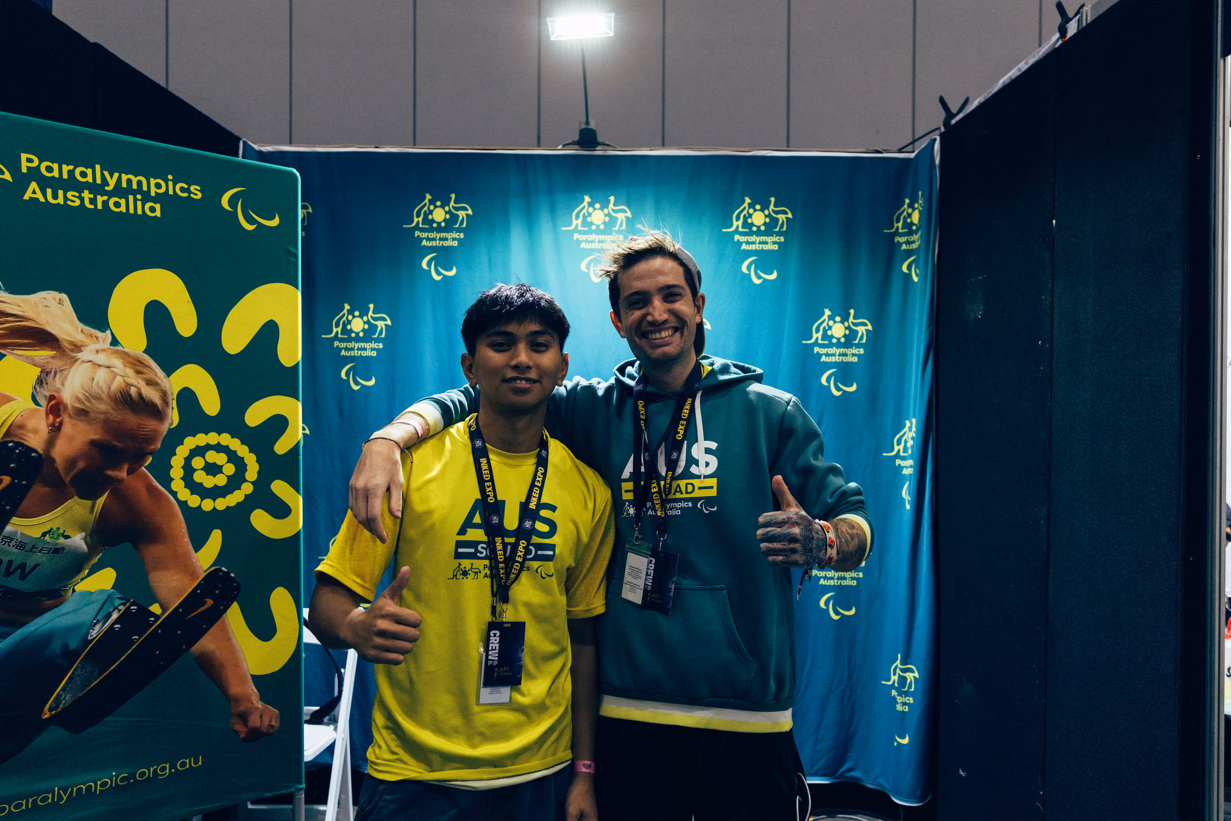 Two young men smiling and giving thumbs up in front of a Paralympics Australia banner, one wearing a yellow shirt and the other a teal hoodie.