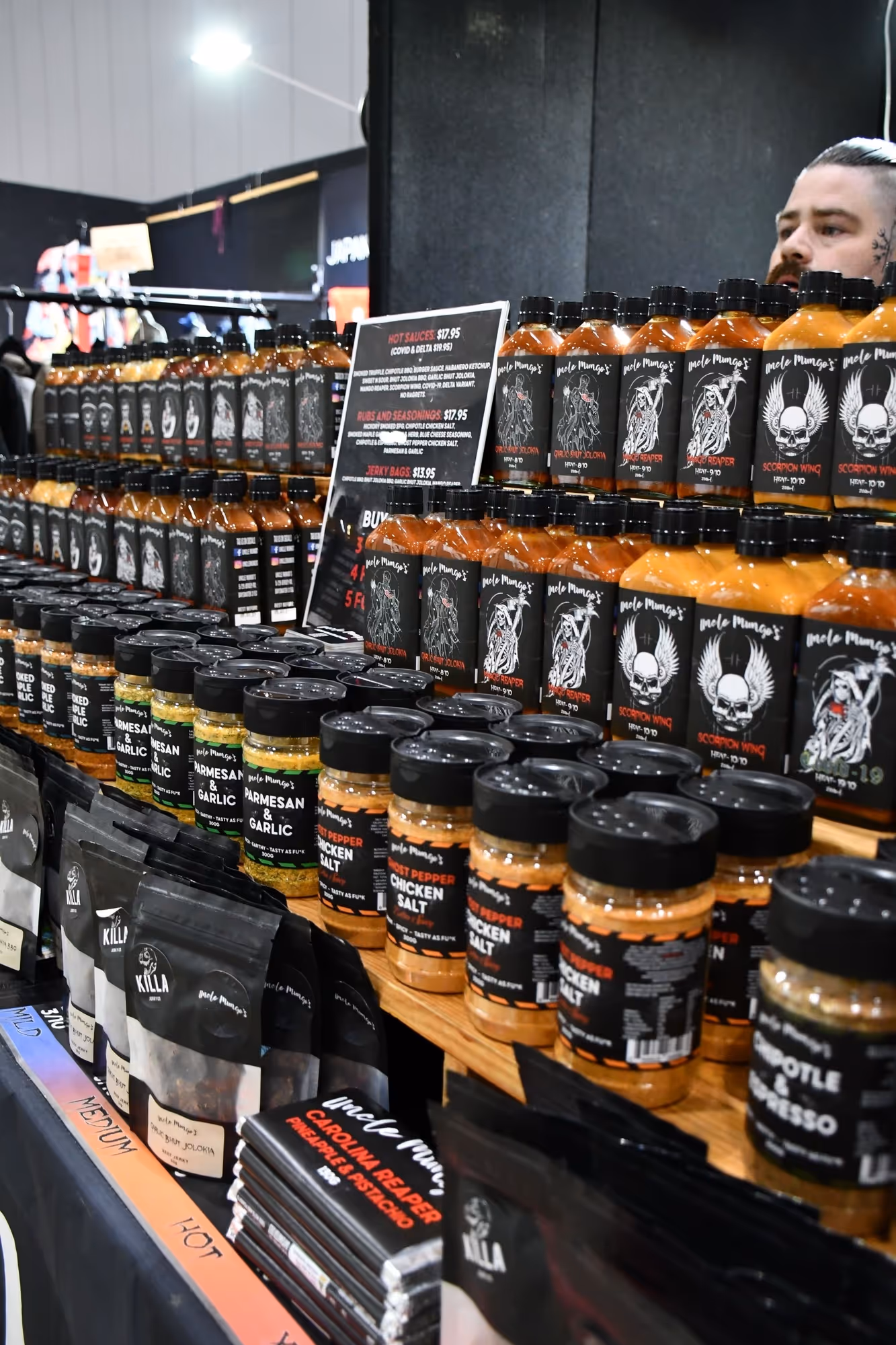 Display of various hot sauces, seasonings, and jerky bags on wooden shelves at a market booth with a person visible in the background.