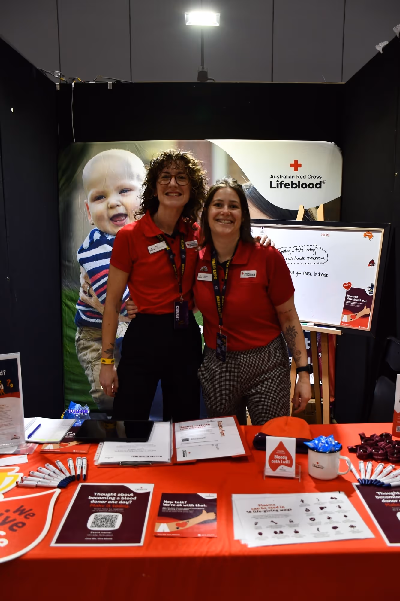 Two smiling women wearing red Australian Red Cross Lifeblood shirts standing behind a red information table promoting blood donation.