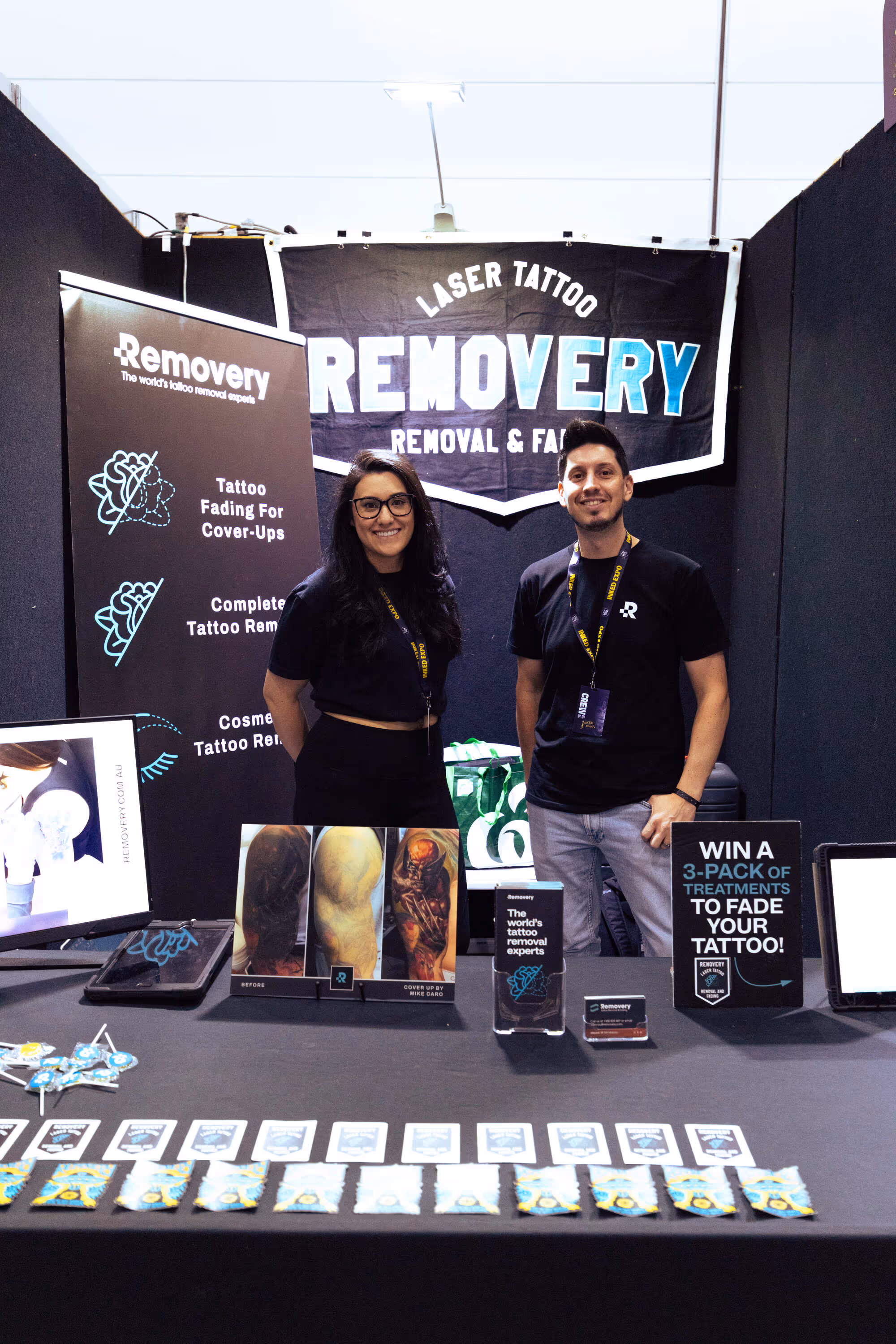 Two smiling people standing behind a booth for Removery laser tattoo removal at an event, with promotional materials displayed on the table.