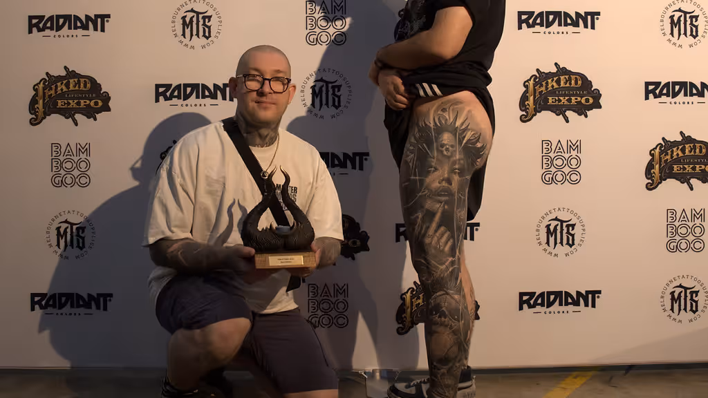Tattooed man with glasses holding a trophy kneeling next to a person showing a large detailed tattoo on their thigh at a tattoo expo.