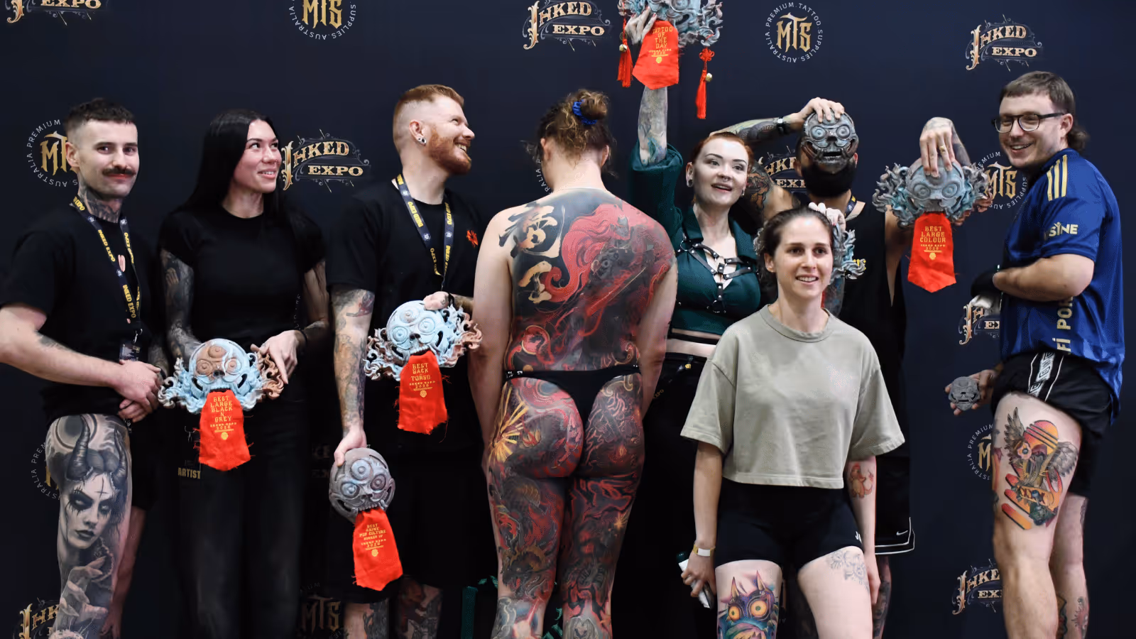 Group of tattoo artists and a model with full back tattoo posing with their award plaques at a tattoo expo.