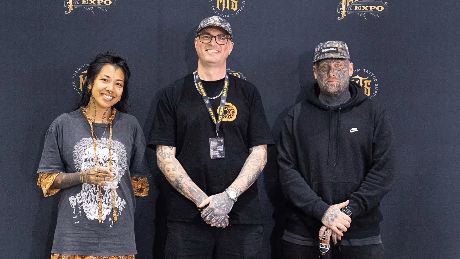 Three people with tattoos smiling and standing against a dark backdrop with expo logos.