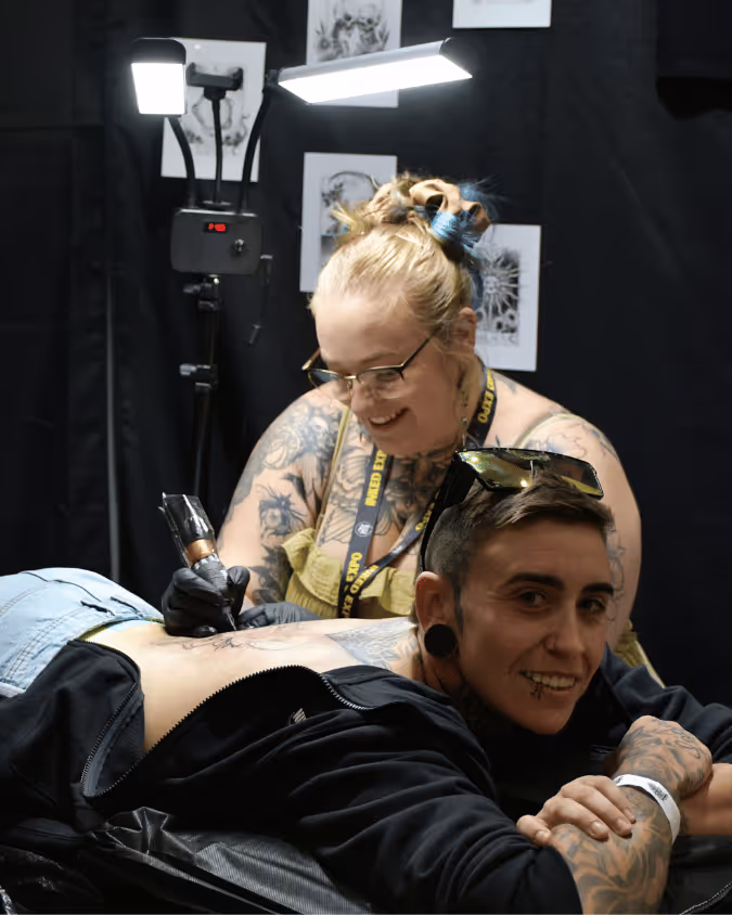 Tattoo artist with glasses and a floral tattoo sleeve working on a tattoo on a smiling person's side, under bright lights.