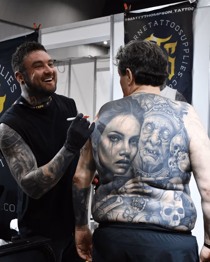 Tattoo artist smiling while working on a large detailed black and gray back tattoo depicting a woman, a skull, and a grotesque crowned figure with skeletal hands on an older man's back.