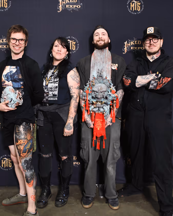 Four tattoo artists with multiple tattoos posing in front of a Inked Expo backdrop, two holding ornate sculptural pieces with a skull design and red tassels.