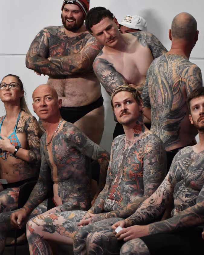 Group of tattooed people sitting and standing indoors, mostly shirtless, showing extensive body art.