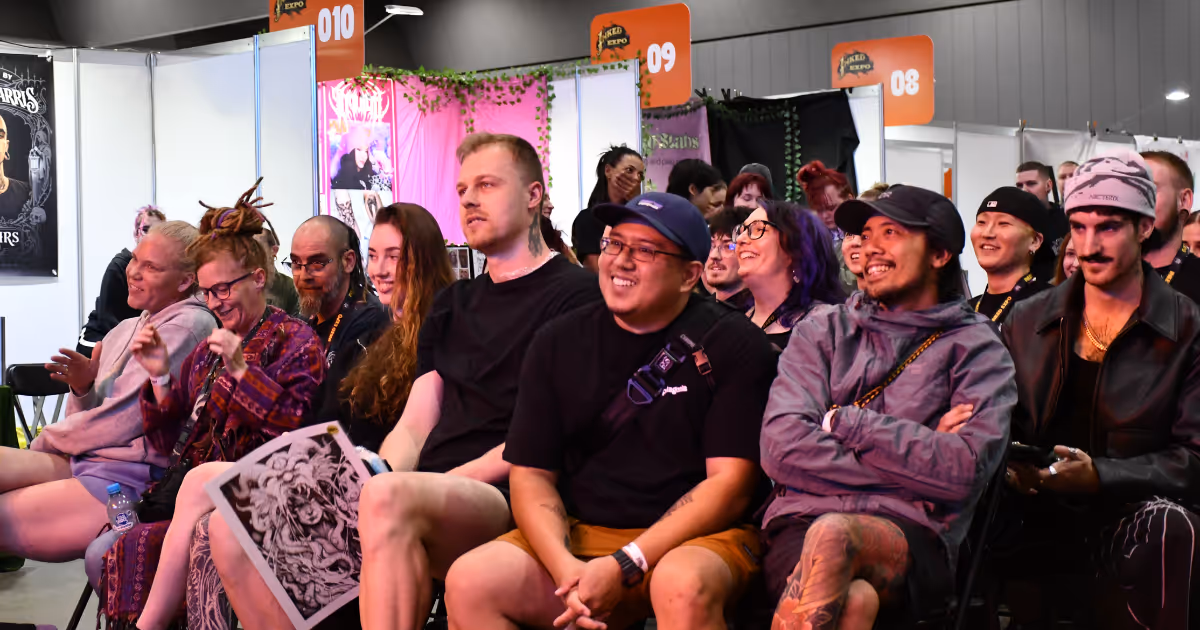 A diverse group of people sitting and smiling at an indoor event with booths and numbered signs in the background.