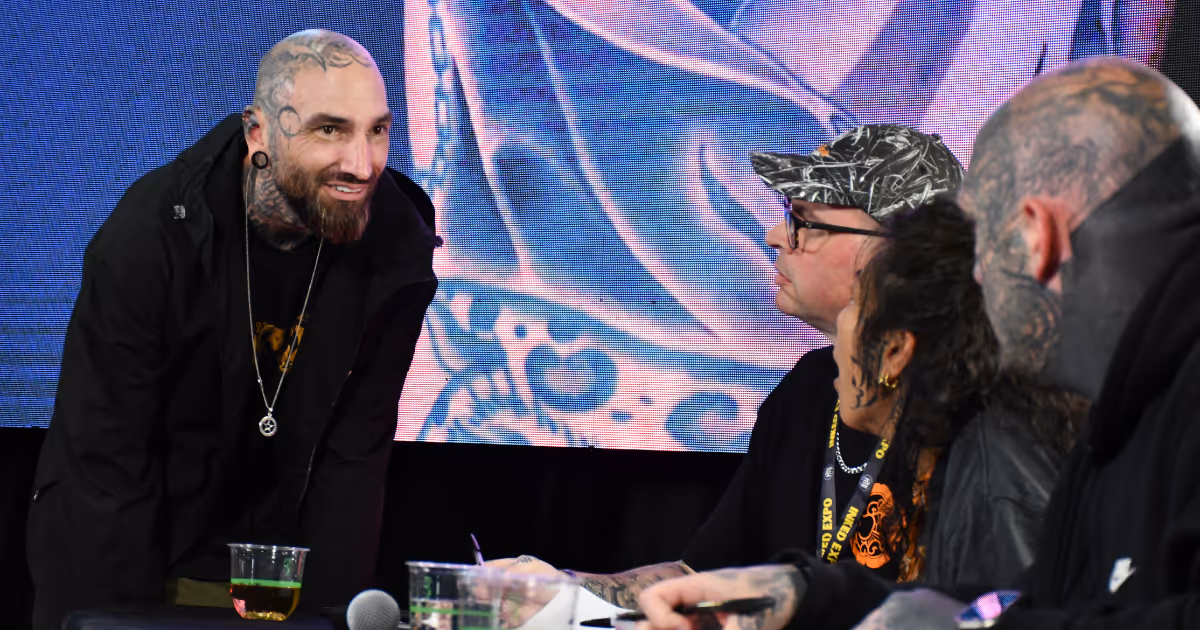 Tattooed man with a beard leaning forward and smiling while talking to three people seated at a table with drinks and microphones.