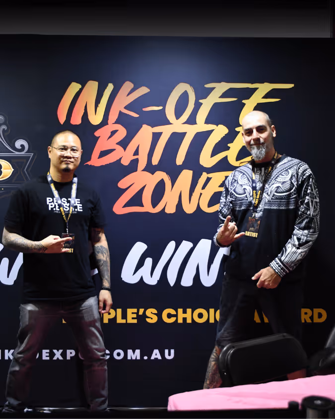 Two men posing with tattoos in front of a sign that reads 'Ink-Off Battle Zone' during a tattoo event.
