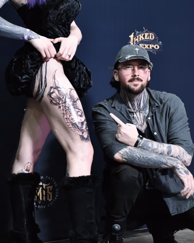 A tattoo artist with sleeve tattoos and facial tattoos crouches next to a person showing a large detailed tattoo on their thigh at a tattoo expo.