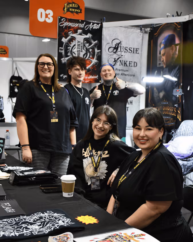 Group of five people at a tattoo expo booth smiling, with tattoo-themed banners and artwork on the table.