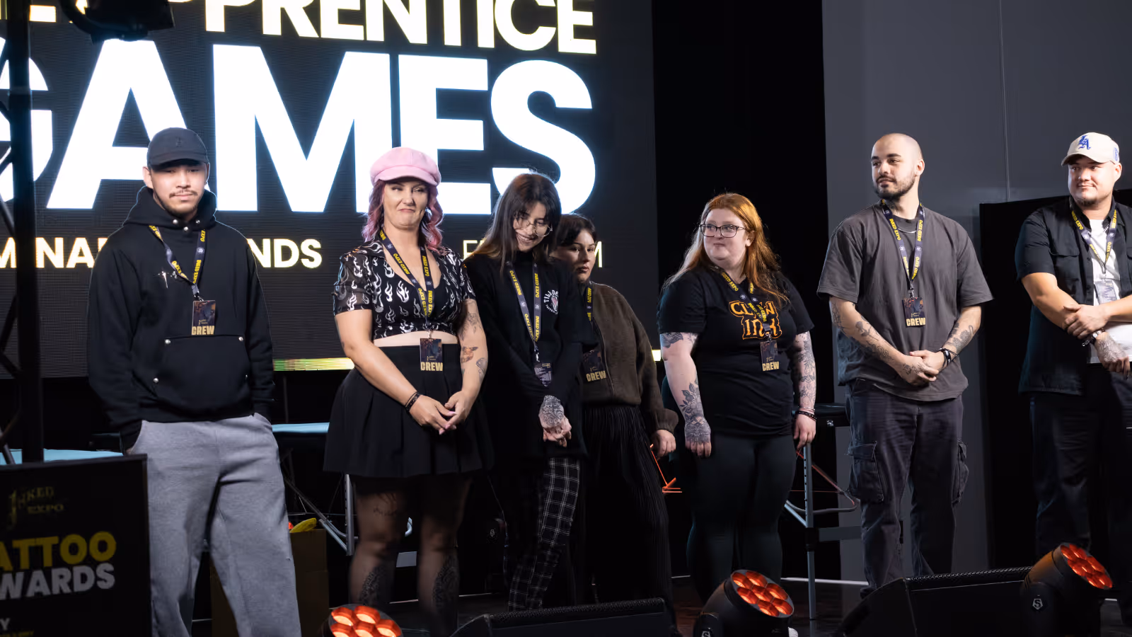 Six people standing on stage with event crew badges and visible tattoos, against a backdrop with large illuminated text.