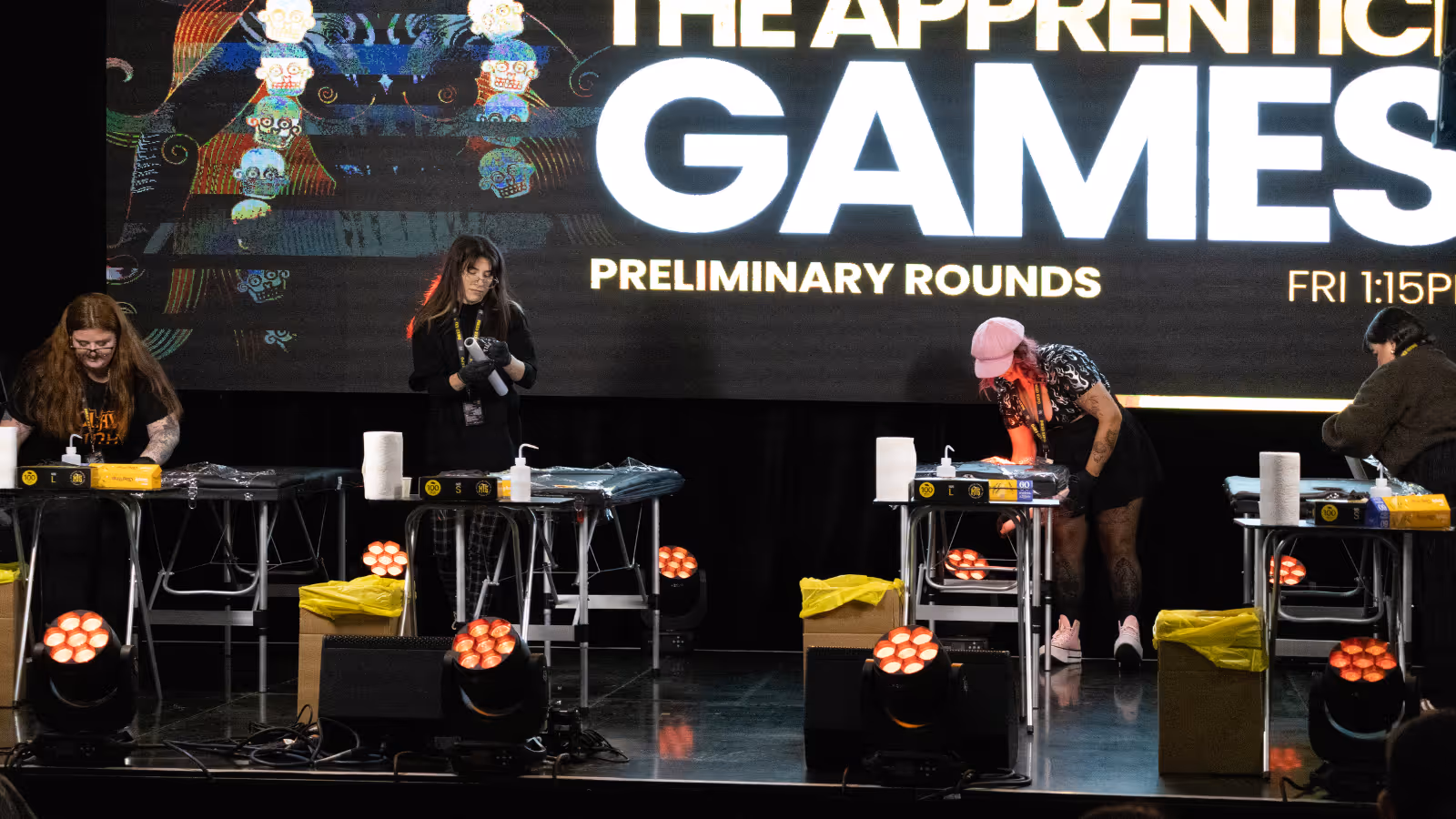 Four people working on separate tattoo stations during The Apprentice Games preliminary rounds event on stage.