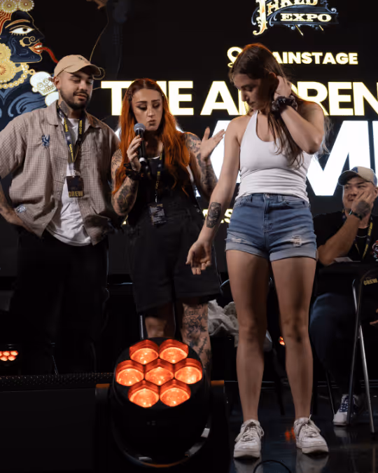 Three people on stage at a convention, one woman with red hair holding a microphone and gesturing, another woman in shorts looking down, and a man in a cap and badge standing beside them.