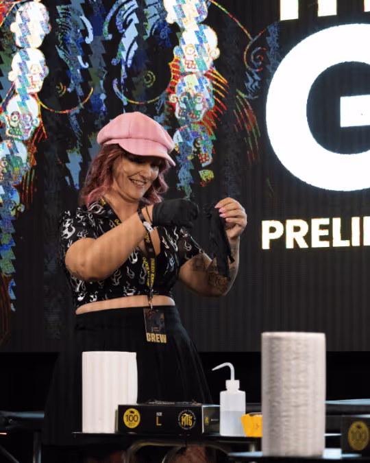 Woman wearing a pink hat and black flame-patterned top smiling while putting on black gloves on stage with various supplies on a table in front of her.