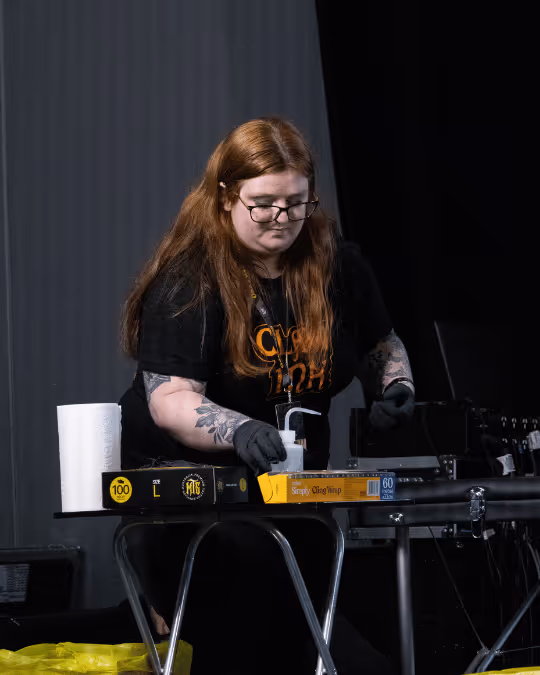 Woman with long red hair, glasses, and black gloves handling a hand sanitizer dispenser on a table with plastic wrap and paper towels.