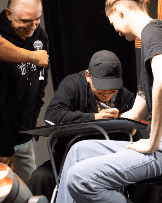 A man in a black cap signing an autograph for a seated person, while another man nearby holds a microphone and smiles.
