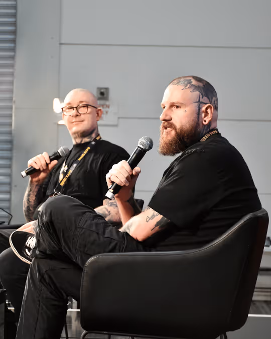 Two tattooed men in black clothing sitting on chairs and speaking into microphones at an indoor event.