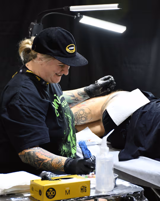 Tattoo artist wearing black gloves and a cap, smiling while working on a tattoo on a client's thigh under bright adjustable lights.