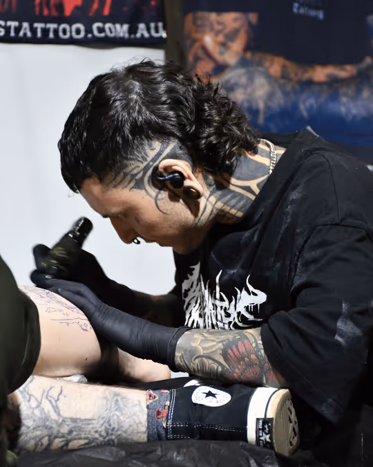 Tattoo artist with black hair and multiple tattoos concentrating on tattooing a client’s leg.
