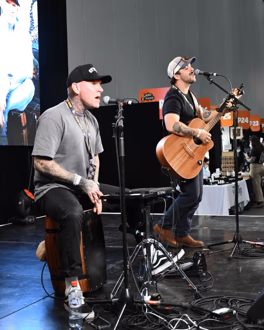 Two tattooed musicians performing on stage, one playing a cajón drum and the other playing an acoustic guitar, both singing into microphones.