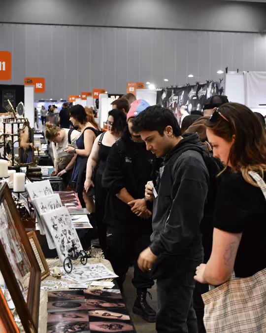 People browsing artwork and prints displayed on tables at an indoor market or convention.