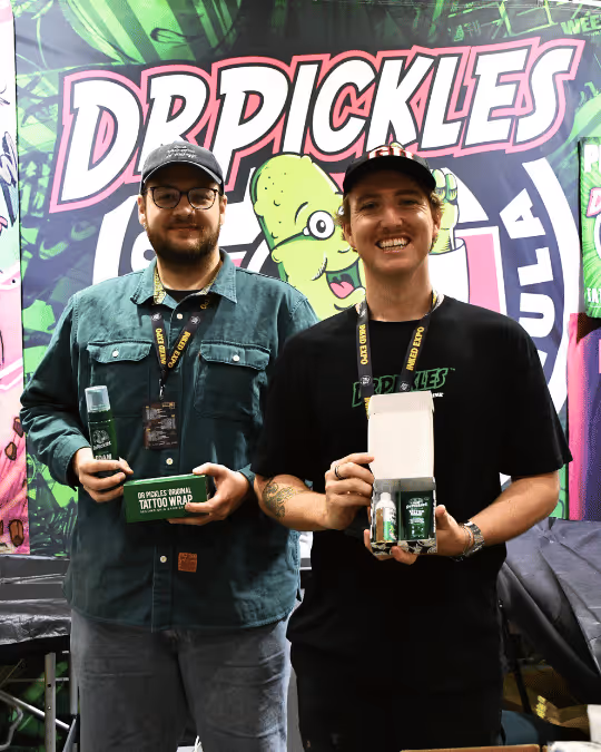 Two men smiling and holding Dr. Pickles tattoo wrap products in front of a branded Dr. Pickles backdrop at a trade show booth.
