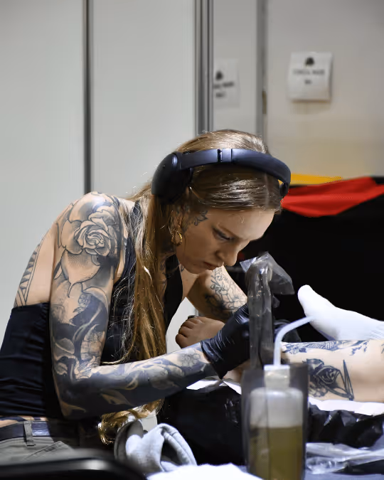 Tattoo artist with floral sleeve tattoos and headphones intently working on a client's leg tattoo.