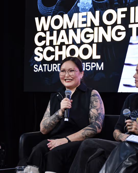 Woman with tattoos and glasses speaking into a microphone in front of a screen that says 'Women of I Changing T School Saturday 15PM'.