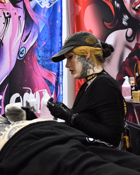 Tattoo artist with black gloves and cap working on a client's leg tattoo in a colorful studio.