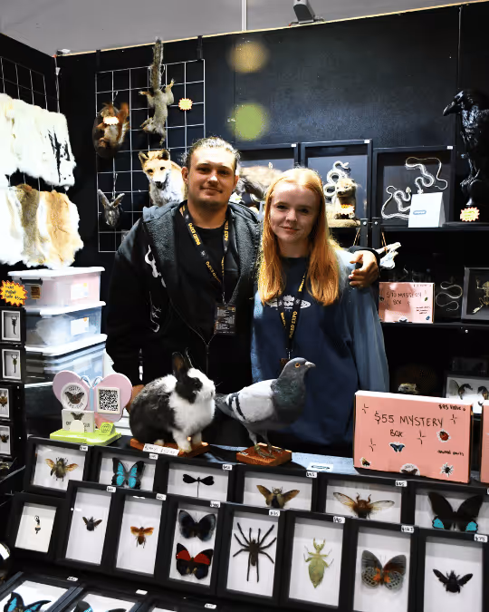 Two people standing behind a display booth with taxidermy animals, mounted insects, and a rabbit and pigeon models on the table.