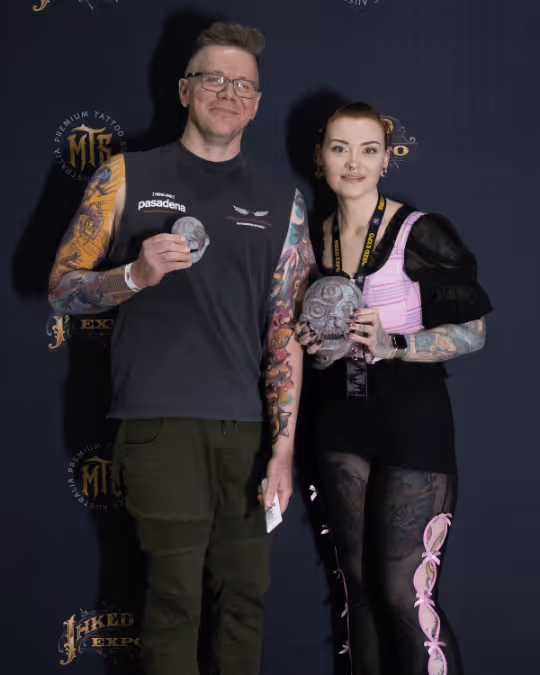 Man and woman with colorful tattoos posing together, each holding a skull sculpture against a dark background with tattoo expo logos.