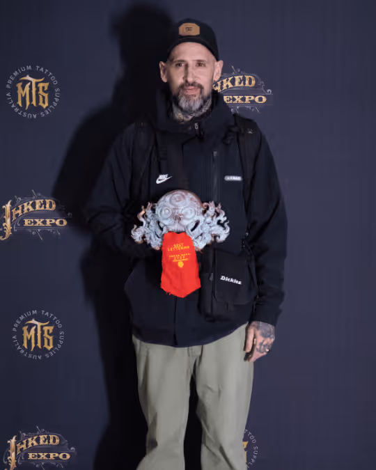 Man wearing a black jacket and cap holding a detailed octopus-shaped trophy with a red ribbon at the Inked Expo event backdrop.