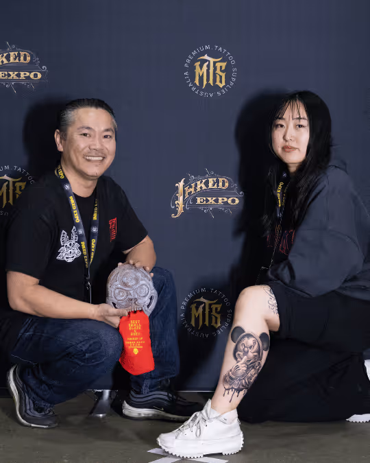 Two people posing in front of a backdrop with 'INKED EXPO' and 'MTS Premium Tattoo Supplies' logos, one showing a detailed tattoo on their leg and the other holding a carved skull trophy with a red ribbon.
