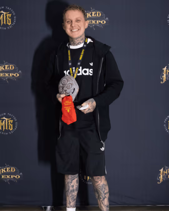 Smiling man with tattoos wearing black Adidas outfit, holding a decorative award and a red cloth, standing in front of a Wicked Expo backdrop.