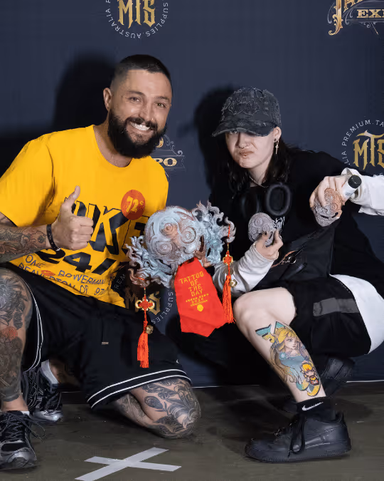 Two tattooed individuals crouching and holding a decorative mask with 'Tattoo of the Day' printed on a red cloth attached to it.