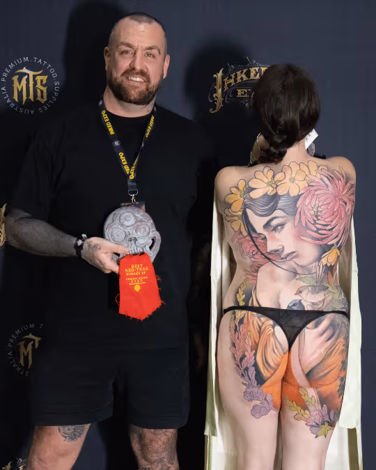 Man in black shirt and shorts holding an award stands next to a woman showing a colorful full-back tattoo of a woman's face with flowers and birds.